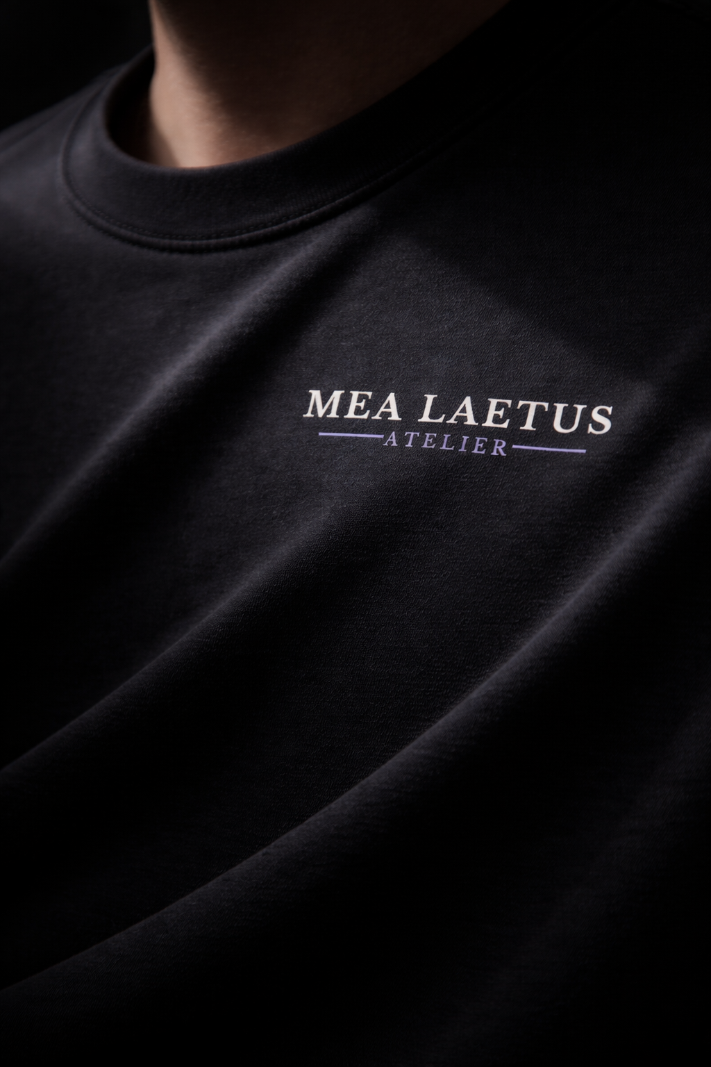 Brustlogo Detailshot von schwarzem Atelier Signature T-Shirt der Fashionbrand MEA LAETUS bei dem ein Lichtstrahl auf das Brustlogo fällt um es in Szene zu setzen.