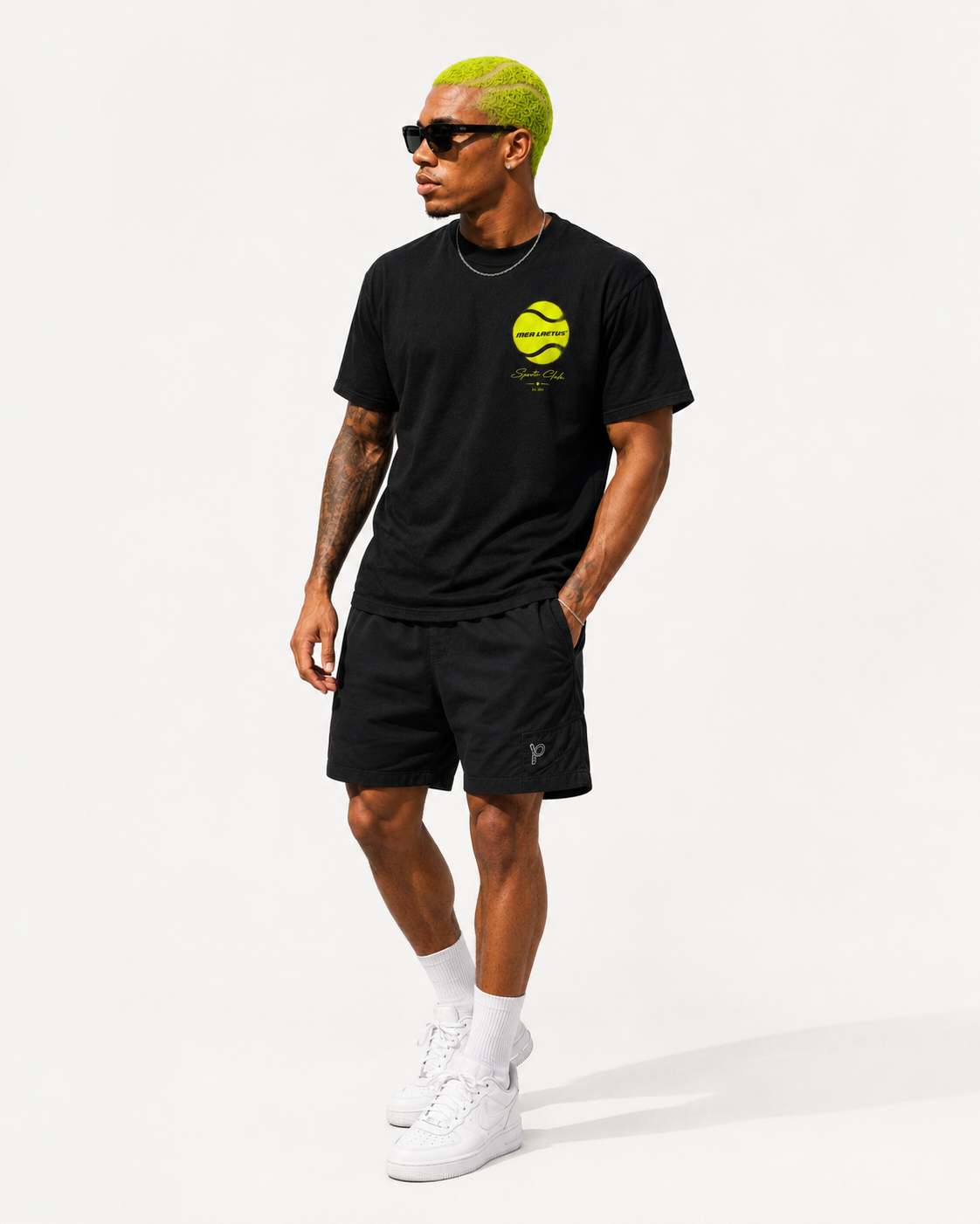 Ganzkörperaufnahme eines Models im schwarzen Graphic Tee mit Tennisball-Brustmotiv, schwarzen Shorts und weißen Sneakern.
