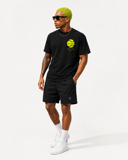 Ganzkörperaufnahme eines Models im schwarzen Graphic Tee mit Tennisball-Brustmotiv, schwarzen Shorts und weißen Sneakern.