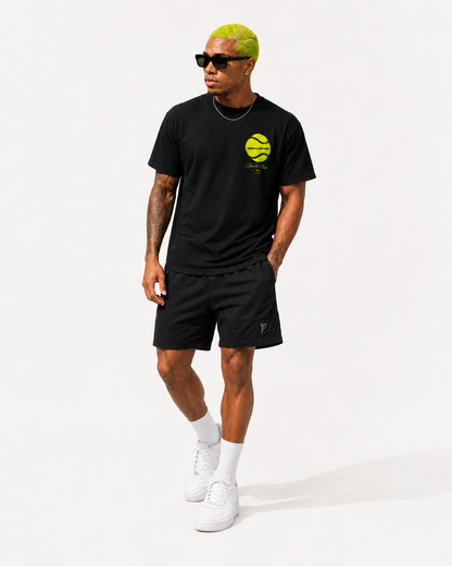 Studioaufnahme eines Models im schwarzen Sportcore-Herrenshirt mit neongelbem Brustmotiv, Sonnenbrille und lässigem Sommer-Outfit.