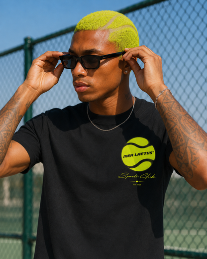 Close-up eines Herren-Shirts mit neongelbem Tennisball-Print vor einem Tennisplatz, getragen mit schwarzer Sonnenbrille.