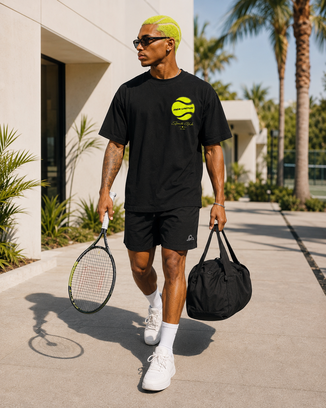 Model auf dem Weg zum Tennisplatz im schwarzen Sports Club Shirt, mit Tennisschläger, Sporttasche, Shorts und weißen Sneakern.