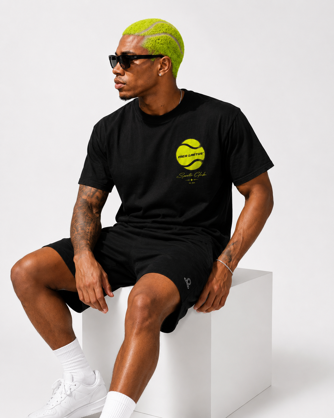 Sitzendes Model im schwarzen MEA LAETUS Tennis Sports Club T-Shirt mit kleinem Neon-Tennisball-Frontprint und schwarzen Shorts.