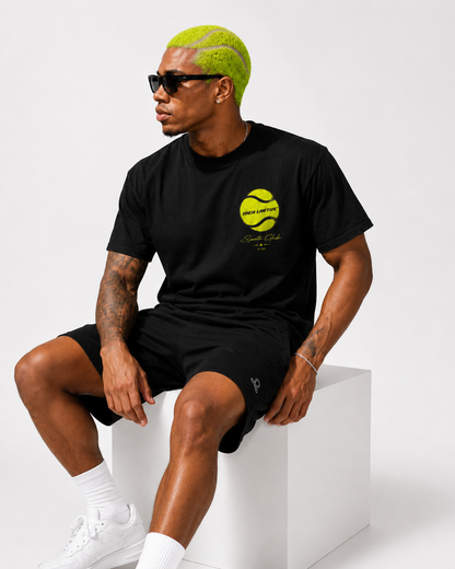 Sitzendes Model im schwarzen MEA LAETUS Tennis Sports Club T-Shirt mit kleinem Neon-Tennisball-Frontprint und schwarzen Shorts.