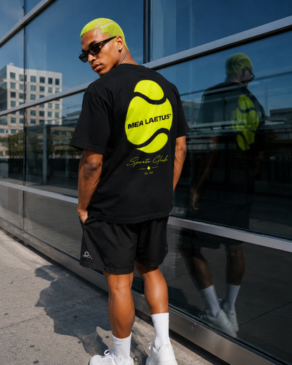 Männliches Model trägt das schwarze MEA LAETUS Tennis Sports Club T-Shirt mit großem Neon-Tennisball-Backprint vor einer Glasfassade.